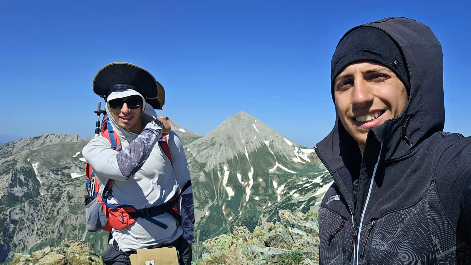 Two hikers on a rocky mountain with pointed peaks behind them