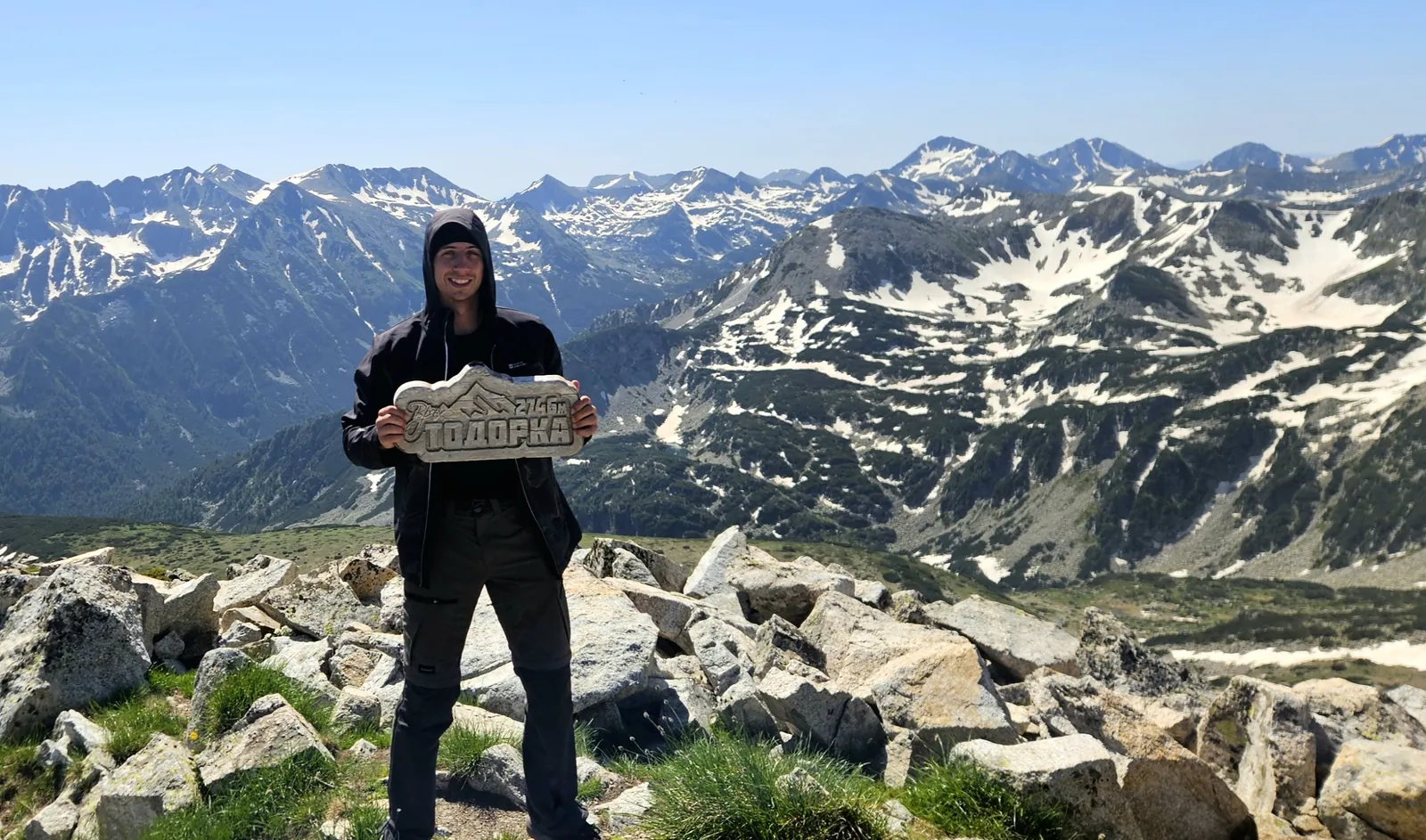 A person holding a sign on a rocky summit above snow-streaked mountains
